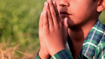 A young holds his hands in prayer while kneeling in a field