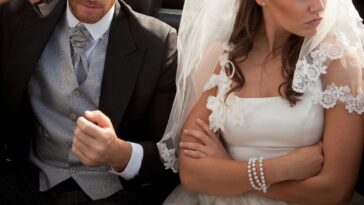 A bride and a groom sit in the backseat of a car, the groom is in an angry conversation on the phone, the bride sits with her arms crossed looking away in anger
