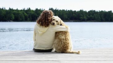 A woman sits on a dock overlooking a lake while hugging her dog