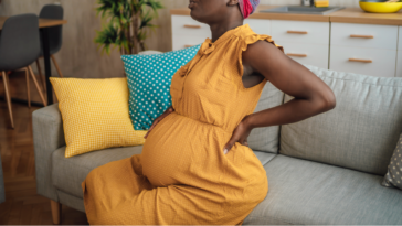 A pregnant woman holding her back sitting on the sofa.