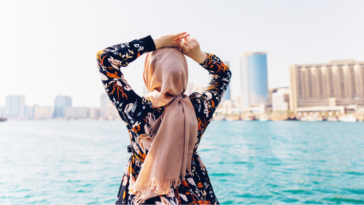 a woman in a hijab looking out at the water with her back to camera.