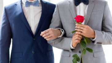 Two men in suits arm in arm. The one on the right holds a rose.