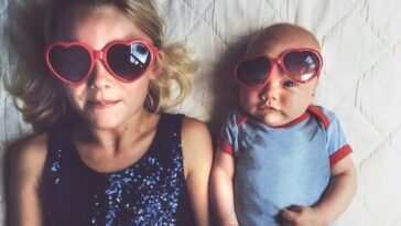 Young girl laying beside baby brother both wearing red love heart glasses