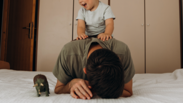 A man lying on a bed while his son climbs on top of him.