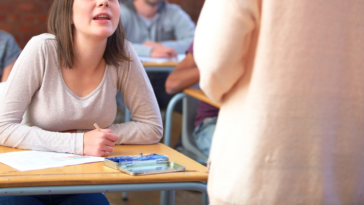 teen girl in high school classroom speaks to another student standing