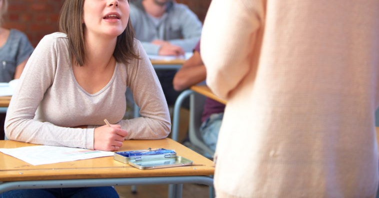 teen girl in high school classroom speaks to another student standing