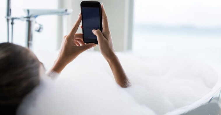 Close up of a woman using cell phone in bubble bath in a bathroom.