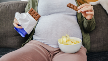 pregnant woman seated on couch eating potato chips and chocolate