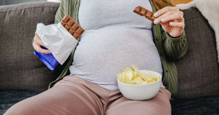 pregnant woman seated on couch eating potato chips and chocolate