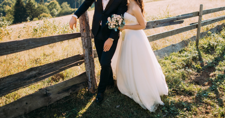 bride and groom standing next to rustic wood fence in field