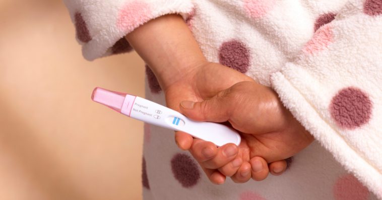 woman in pink bathrobe holding pregnancy test behind her back