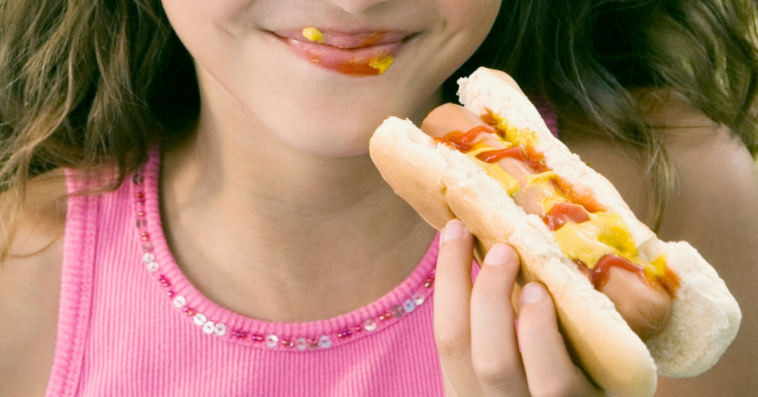 Young girl holding a hot dog with mustard on her face.