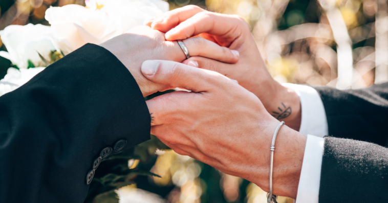 A groom slipping a wedding band on his husband-to-be's finger.