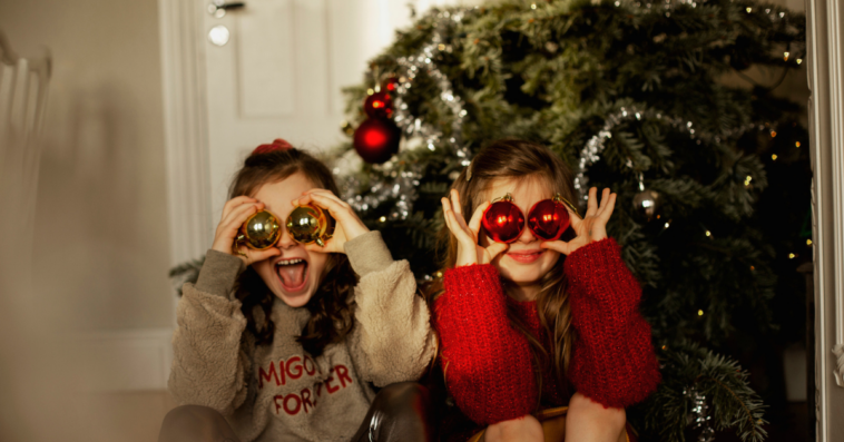 Two children sitting by a Christmas tree, covering their eyes with Christmas ornaments.