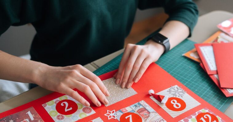 Close-up top view of unrecognizable young woman gluing envelopes on board with gifts for children making Christmas advent calendar at home