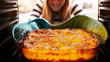 A smiling woman Taking a Cooked Dish Of Lasagne Out Of The Oven.