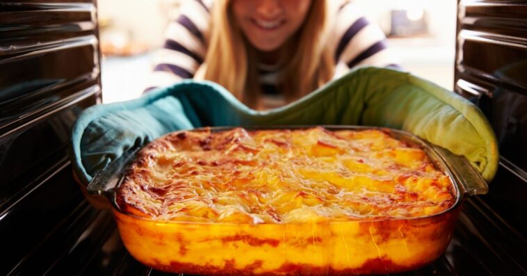 A smiling woman Taking a Cooked Dish Of Lasagne Out Of The Oven.