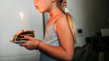 Cute girl with pigtails holds a piece of cake while blowing out candle, children's birthday.