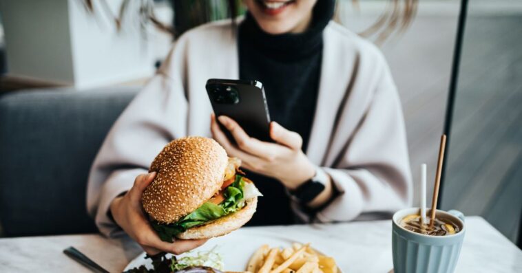 Smiling young woman sitting by the window in a restaurant enjoying her lunch. She is taking photos of her delicious food, fresh burger with fries with smartphone. Eating out lifestyle.