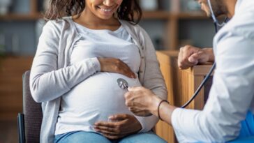 Pregnant millennial woman sitting on chair at clinic, touching her big tummy and smiling, visiting doctor, arab man gynecologist checking baby heart rate with stethoscope.