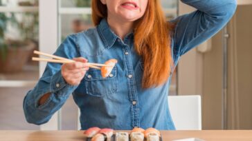 Redhead woman eating sushi using chopsticks stressed with hand on head, shocked with shame and surprise face, angry and frustrated.