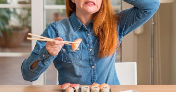 Redhead woman eating sushi using chopsticks stressed with hand on head, shocked with shame and surprise face, angry and frustrated.