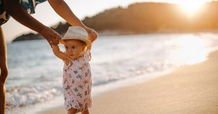 Mom walks baby girl on a beach, at sunset.