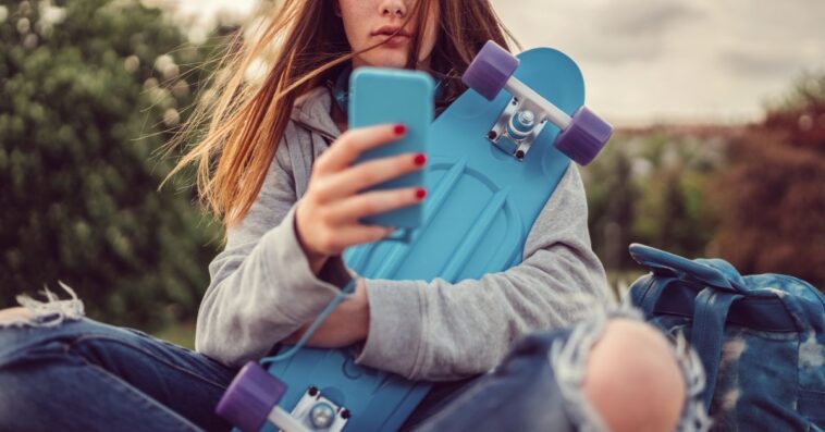 Teenage girl with skateboard texting on smartphone.
