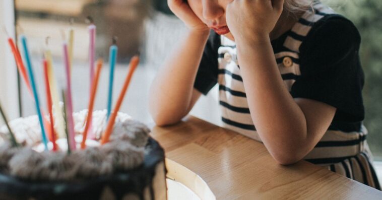 Cute young girl wearing a party hat, sulking in front of a birthday cake.