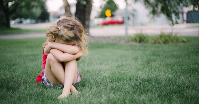 Little girl with curly hair sits on grass and cries into her arms.