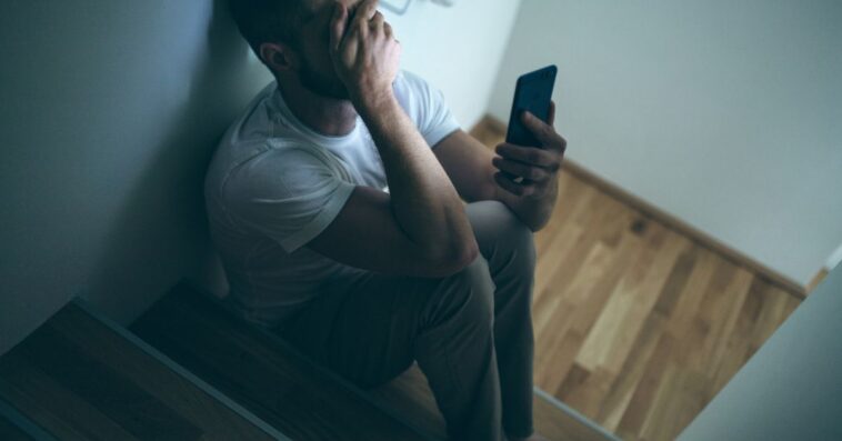 Depressed man sitting in the dark with his smartphone.