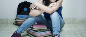 Indoor shoot of a high school teenage girl student sitting on floor with books, bag and mobile phone in her hand. She is in blue top, jeans and purple canvas shoes