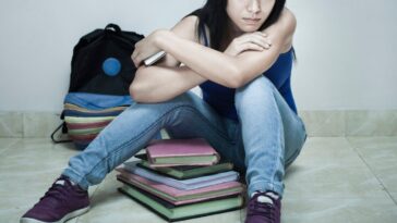 Indoor shoot of a high school teenage girl student sitting on floor with books, bag and mobile phone in her hand. She is in blue top, jeans and purple canvas shoes