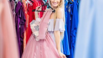 Beautiful woman shopping and choosing the right dress for luxury event. Young woman holding and trying on elegant gown and smiling at camera.