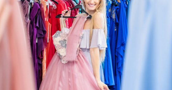 Beautiful woman shopping and choosing the right dress for luxury event. Young woman holding and trying on elegant gown and smiling at camera.