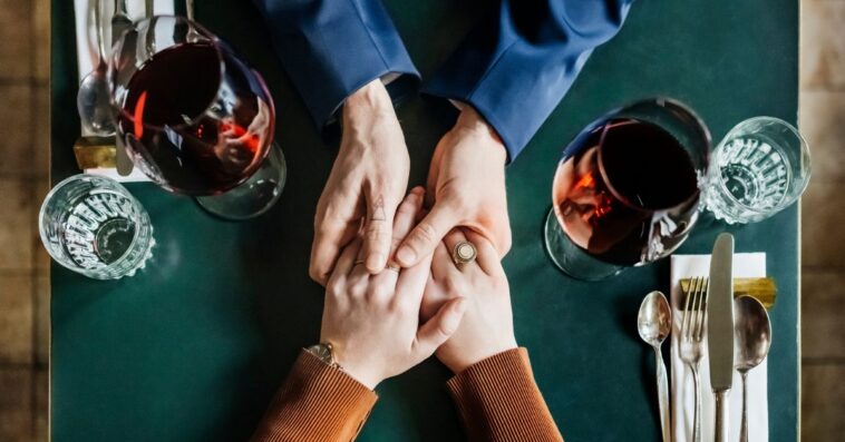 An aerial view of a couple holding hands and drinking red wine while sitting at a restaurant table for lunch together.