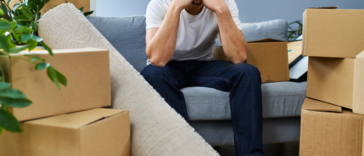 unhappy young man seated on couch surrounded by moving boxes