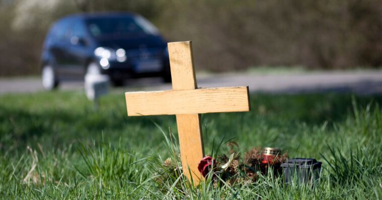 A wooden roadside memorial. In the background an unrecognisable car drives by the scenery.