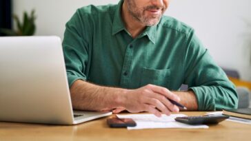 Man at home sitting with laptop, calculator and documents examining home finances.