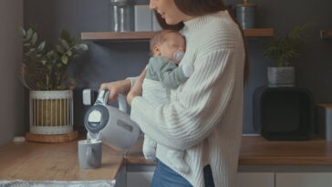 Young woman carrying baby while pouring herself cup of tea