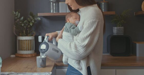 Young woman carrying baby while pouring herself cup of tea