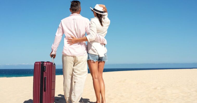 Happy traveling couple standing with suitcases on a sandy beach on a sunny summer day.