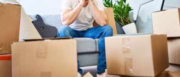 Frustrated man sitting on couch surrounded by cardboard boxes and miscellaneous.