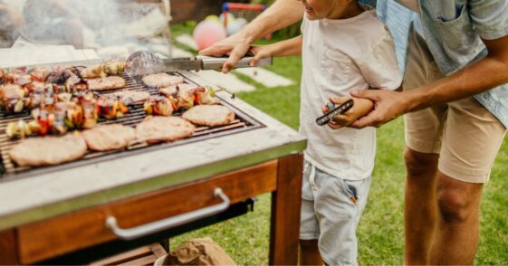Photo of father and son grilling meat during a barbecue party in their yard.