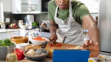 Man in his 20s making lunch, cutting bell peppers, using digital tablet with online recipe in kitchen, enjoyment, connection, domestic life.