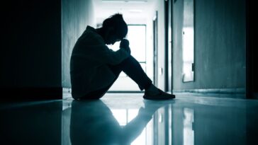 A young woman sits in a dark hallway, hanging her head in her hands.