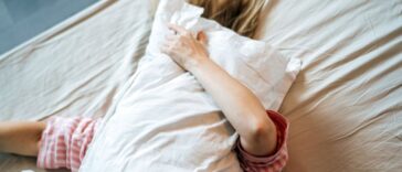 A woman lies in bed with a pillow in her hands, covering her face with it, her hair spread over white sheets.