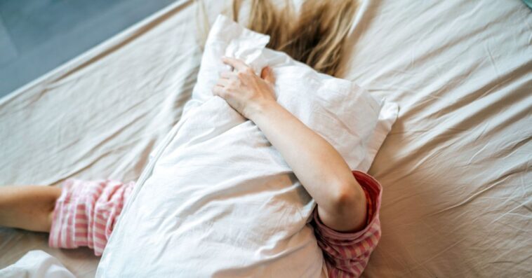 A woman lies in bed with a pillow in her hands, covering her face with it, her hair spread over white sheets.