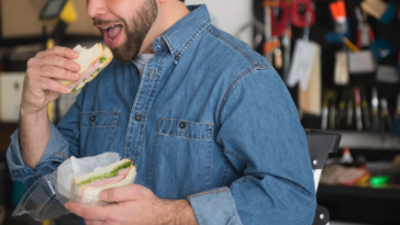 A man in a denim shirt eating a sandwich, with tools in the background.