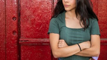 Young woman, arms crossed and looking impatient against a red door.